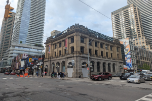 Eine belebte Stadtstraße mit hohen Gebäuden, Fahrzeugen, Fußgängern, Verkehrsampeln, Bäumen und der Toronto Public Library als Mittelpunkt unter einem bewölkten Himmel.