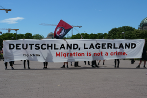 Eine Gruppe von Menschen, die auf der Straße stehen und eine Fahne halten, auf der steht "Deutschland, Lagerland Migration ist kein Verbrechen" und eine Fahne, mit Straßenlaternen, Bäumen, Kränen, einem Gebäude und einem bewölkten Himmel im Hintergrund.