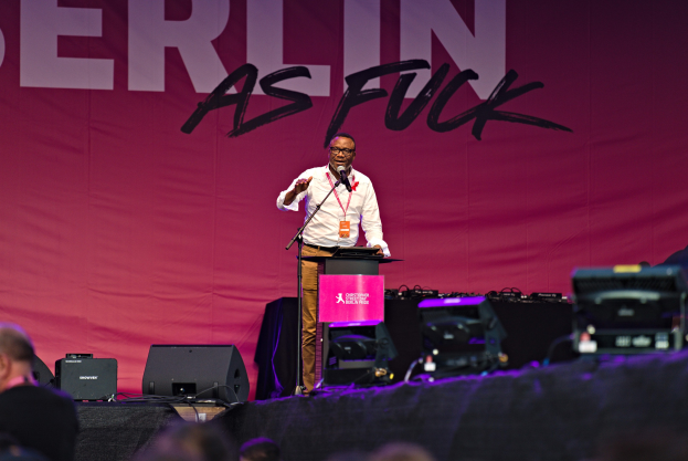 Ein Mann in weißem Hemd und braunen Hosen steht auf einer Bühne und hält ein Mikrofon in der Hand, hinter ihm eine pinkfarbene Flagge mit der Aufschrift 'Berlin as Fuck' und sichtbare Lautsprecher.