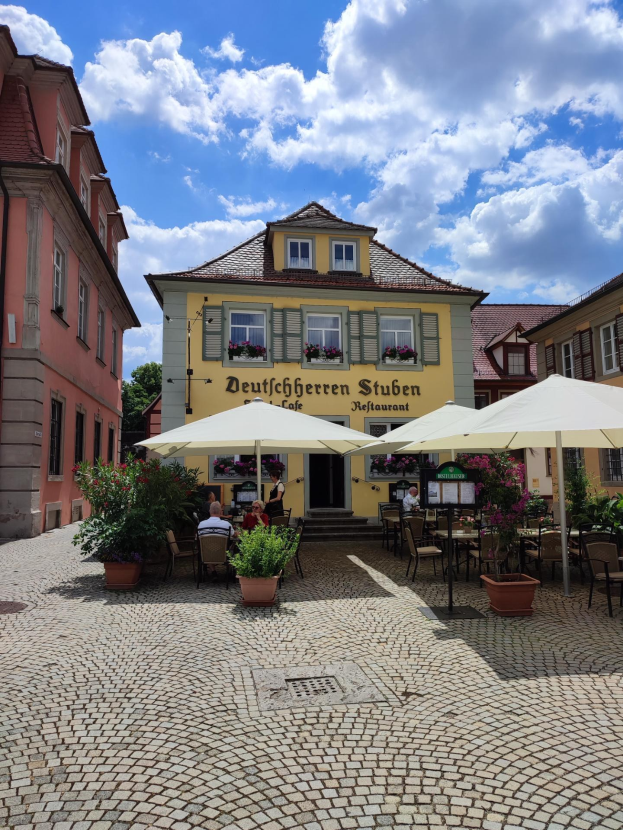 Außensitzbereich des Deutschen Stuben Restaurants in Baden-Württemberg, Deutschland, mit Kopfsteinpflasterstraße, gelbem Gebäude, Tischen, Sonnenschirmen und Menschen.