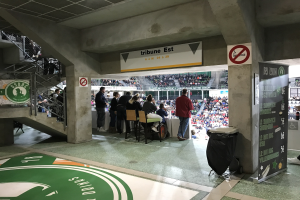 Gruppe von Menschen, die vor einer sitzenden Menge in einem Stadion mit Schildern, einem Mülleimer, einer Tafel, einer Treppe und einem Dach mit Lampen stehen; Text auf den Schildern lautet "Boston Celtics vs New York Jets".