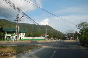 Tankstelle mit Strommasten, Kabeln, Schildern, Bäumen, Gebäuden, Fahrzeugen, Hügeln und bewölktem Himmel im Hintergrund.