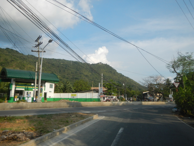 Tankstelle mit Strommasten, Kabeln, Schildern, Bäumen, Gebäuden, Fahrzeugen, Hügeln und bewölktem Himmel im Hintergrund.