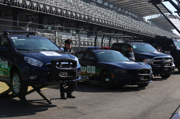 Polizeiautos vor einer Rennstrecke mit uniformierten Personen auf der Straße, Stadion und Absperrungen im Hintergrund.