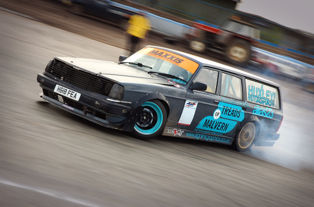 Ein Auto rast eine Rennstrecke entlang und hinterlässt eine Rauchwolke, umgeben von anderen Fahrzeugen und Menschen im Hintergrund.