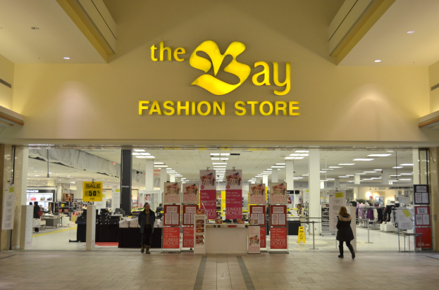 Verkaufsfläche eines Bay Fashion Retail Shop in einem Einkaufszentrum mit vorbeigehenden Kunden, Texttafeln an den Wänden, Deckenleuchten und Säulen im Hintergrund.