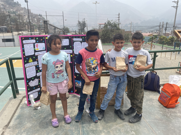 Gruppe von Kindern, die auf einem Basketballfeld stehen und Papiertüten halten, mit Tüten und Gegenständen rechts und einer Tafel mit Text links, vor einer Außenkulisse mit Zaun, Pfosten, Straßenlaternen, Bäumen, Gebäuden, Hügeln und dem Himmel.