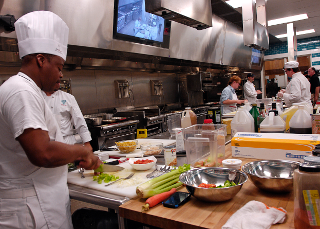 Eine Gruppe von Köchen in einer Küche beim Zubereiten von Speisen auf einem Schneidbrett, wobei ein Koch im Vordergrund einen weißen Kittel und eine Haube trägt und ein Messer hält und von verschiedenen Utensilien, Schüsseln, Gemüse, einem Mobiltelefon, Flaschen und anderen Gegenständen umgeben ist, mit einem Fernseher und Deckenleuchten im Hintergrund.