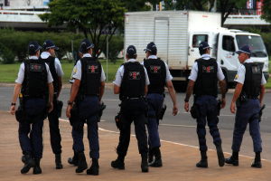 Eine Gruppe von Beamten in Uniform, einschließlich M√ľtzen, Hemden, kugelsicheren Westen, Uhren, Hosen und Schuhen, die auf einer Stra√üe mit einem geparkten Lastwagen auf der rechten Seite n√§her an einem Zaun, Geb√§uden, B√§umen, Pflanzen, Masten und Bannern im Hintergrund und einer Br√ľcke oben rechts gehen.