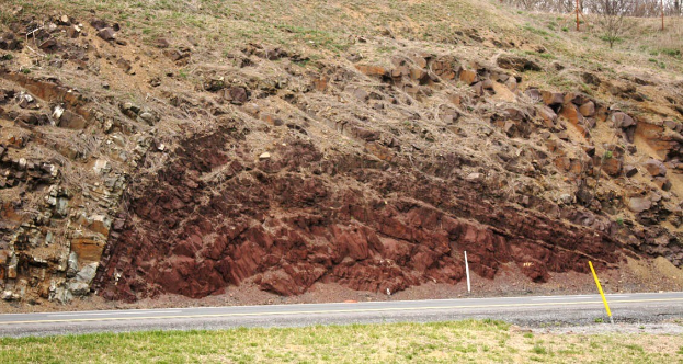 Großer Erdrutsch an der Seite einer Straße mit Gras unten, einem Hügel in der Mitte, Bäumen und Himmel im Hintergrund.