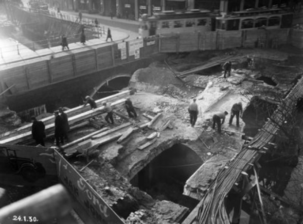Schwarz-weiß-Foto von Bauarbeitern bei der Reparatur einer Brücke, mit Menschen auf der Brücke und Fahrzeugen auf der Straße im Hintergrund.
