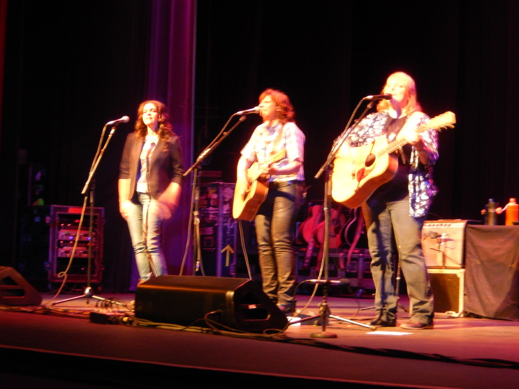 Drei Frauen treten auf der Bühne eines Musikkonzerts auf, jede singt in ein Mikrofon, während sie eine Gitarre spielt, mit einem Tisch voller Wasserflaschen und Gitarren dahinter.