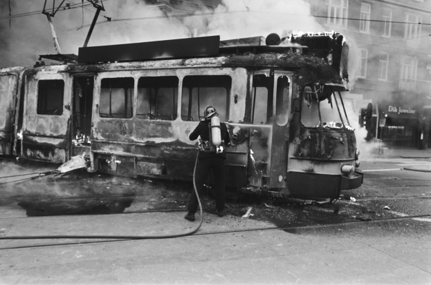 Schwarzes und wei├čes Bild eines Mannes in einem Helm, der eine Pfeife h├Ąlt und neben einer ausgebrannten Stra├čenbahn mit aufsteigendem Rauch steht, auf einer Stra├če mit einem Geb├Ąude und Kabeln im Hintergrund.