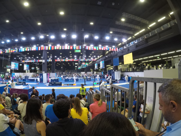 Eine große Menge sitzt und steht in einer Indoor-Sportarena mit Bildschirmen, Fahnen und Deckenleuchten im Hintergrund.