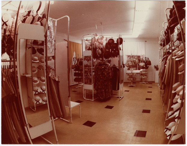 Altes Foto eines Bekleidungsgeschäftsinneren mit Kleidung an Bügeln, Hüten auf Ständern, einem Spiegel, einem Stuhl, einem Tisch mit Gegenständen, Vorhängen und Deckenleuchten.