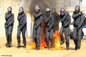 Menschen in Helmen vor einem Feuer mit verschiedenen Gegenst√§nden auf dem Boden, Geb√§ude im Hintergrund, ein Fahrzeug und ein Plakat und eine Tafel an der linken Wand, mit Text am unteren Bildrand.