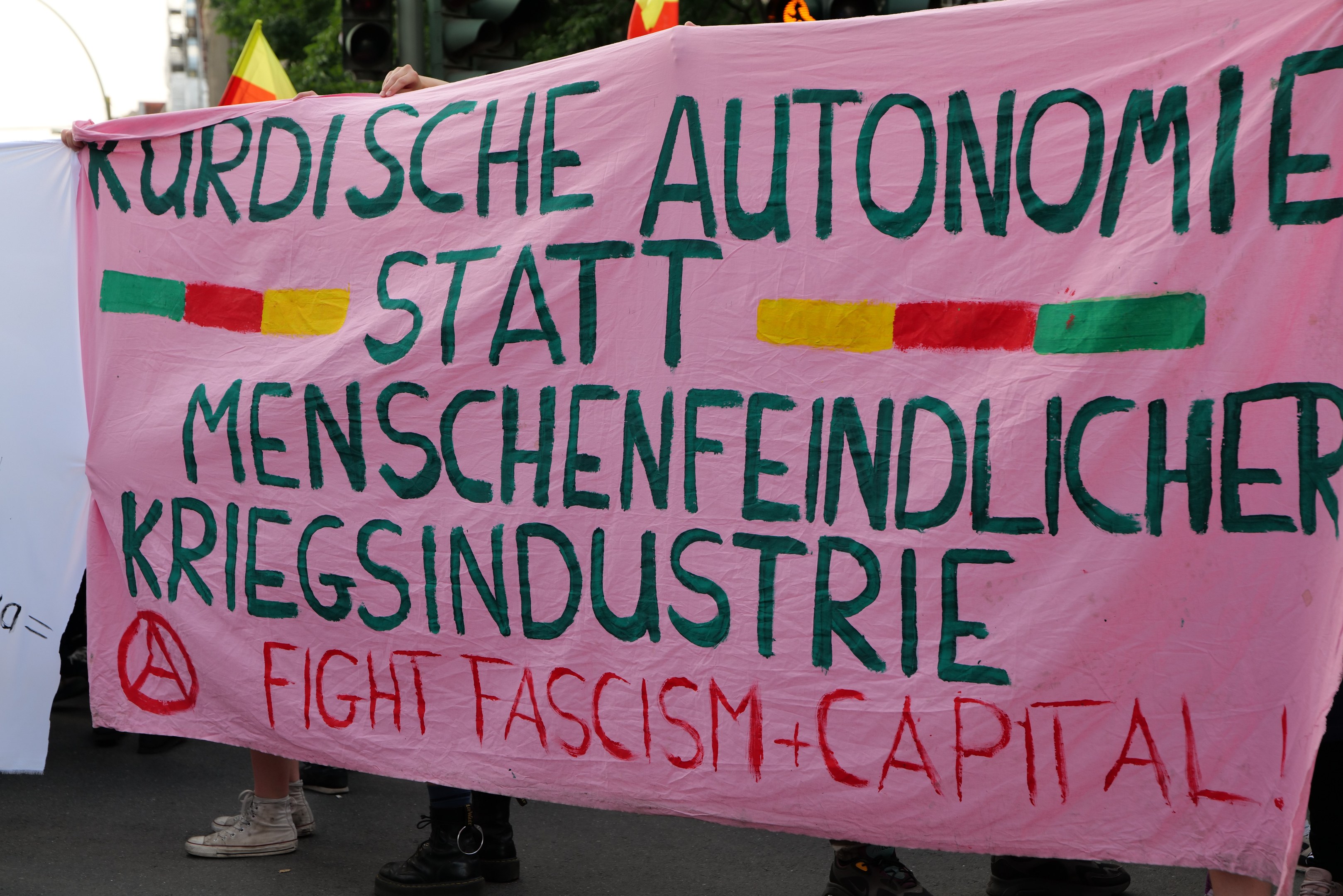 Gruppe von Menschen mit einer pinken Fahne, auf der 'Kurdische Autonomie Statt Menschenfeindlicher Kriegsindustrie Fight Fascism Capital' steht, auf einer Straße mit Ampeln, Flaggen, Bäumen, Gebäuden und einem klaren blauen Himmel im Hintergrund.