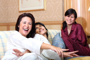 Drei Frauen lachen zusammen, mit einem Kissen neben der mittleren Frau und einem gerahmten Bild an der Wand im Hintergrund.