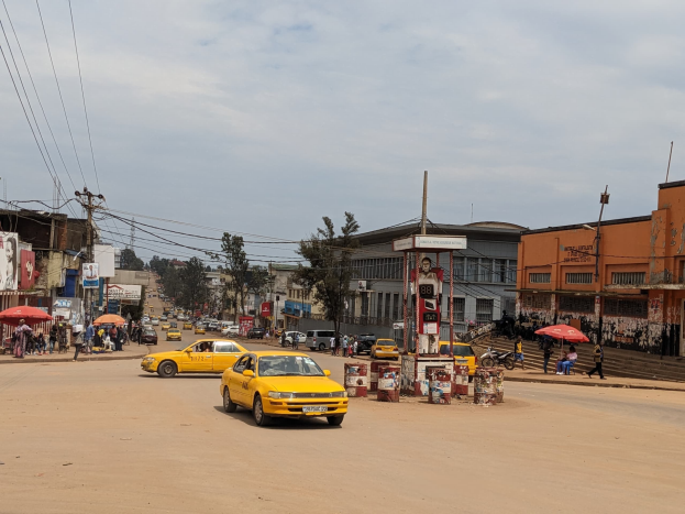 Ein gelbes Taxi fährt eine belebte Stadtstraße mit Gebäuden an den Seiten hinunter, während Fußgänger mit Schirmen auf dem Gehweg darunter gehen und Wolken am Himmel und Strommasten mit Drähten zu sehen sind.