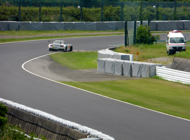 Ein Rennwagen fährt auf einer Rennstrecke mit einem Polizeiwagen im Hintergrund, umgeben von Grünflächen und Absperrungen, mit Text unten.