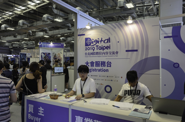 Menschengruppe um einen Stand auf der Digital 2019 Taipei International Digital Content Summit & Fair, mit zwei sitzenden Personen vor einem Tisch mit Papieren, Stiften, einem Laptop, einer Flasche, einem Korb und anderen Gegenständen sowie Bannern mit Text im Hintergrund.