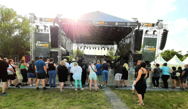 Menschenmenge vor einer Bühne auf einem Musikfestival mit Zelten und Bannern rechts, grasigem Bereich, Bäumen im Hintergrund, bewölktem Himmel mit strahlender Sonne.