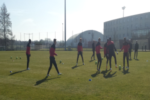 Eine Gruppe von Menschen, die Fußball auf einem Feld spielen, das von einem Zaun, Pfählen, Bäumen, Gebäuden und einem klaren blauen Himmel umgeben ist, mit mehreren Fußballen, die herumliegen, und einigen Spielern, die Schuhe tragen.
