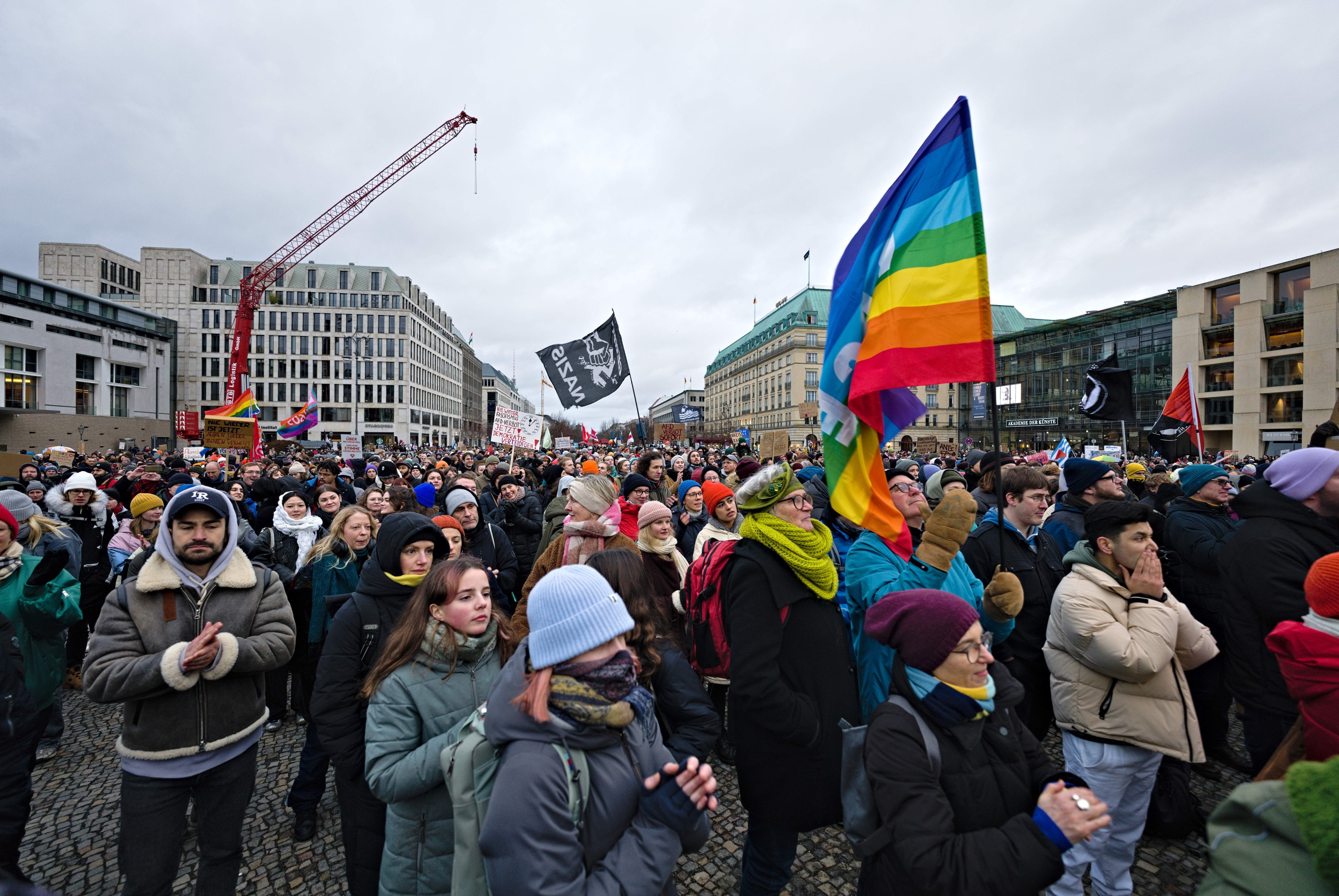 Große Gruppe von Menschen bei einer LGBTQ+-Rechtsdemo in Berlin, die Fahnen und Plakate schwenken, mit Gebäuden, einem Kran und einem bewölkten Himmel im Hintergrund.