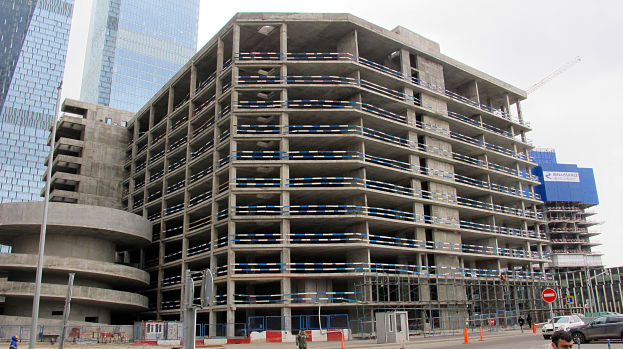 Großer Bau in einer städtischen Umgebung mit Wolkenkratzern, Fahrzeugen, Fußgängern, Verkehrs-Infrastruktur, Kränen und einem klaren Himmel.