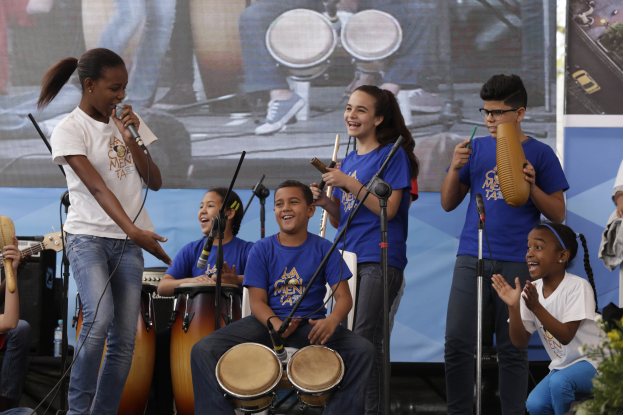 Eine Gruppe von Kindern, die auf einer Bühne Musikinstrumente spielen, mit einer Frau in der Mitte, die ein Mikrofon hält, umgeben von Mikrofonstativen, einem Banner im Hintergrund und Pflanzen auf der rechten Seite.