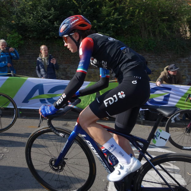 Professionelle Radrennfahrerin Katie Archibald fährt ein Fahrrad die Straße hinunter, trägt einen Helm, eine Schutzbrille, Handschuhe und Schuhe und hat eine Menschenmenge und ein Banner im Hintergrund.