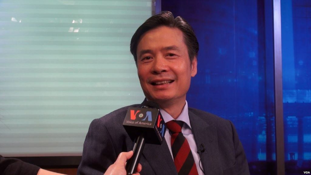 Ein Mann in Anzug und Krawatte hält ein Mikrofon, mit der Hand eines Reporters auf der linken Seite, vor einem Fenster und einer Wand sprechend.