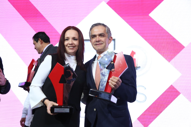 Zwei Personen auf einer Bühne mit Trophäen in den Händen, die lächeln, und einem Awards-Ceremony-Hintergrund.