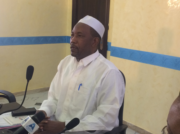 Ein Mann in einem weißen Gewand und einer Mütze sitzt an einem Tisch mit Mikrofonen, während er spricht und Papiere und Gegenstände auf dem Tisch sowie eine Person mit Brille rechts von ihm zu sehen sind, in einem Raum mit einer Wand und einer Tür im Hintergrund.