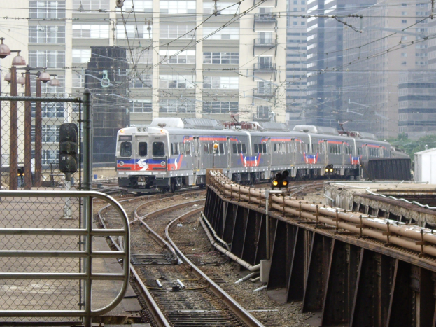Nahverkehrszug fährt auf Schienen neben Hochhäusern mit Bahninfrastruktur und städtischen Elementen im Hintergrund.