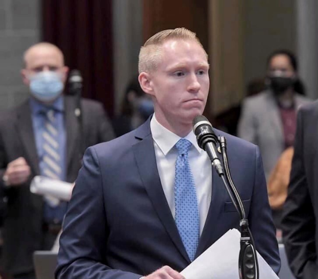 Ein Mann in Anzug und Krawatte spricht an einem Podium mit Papieren vor einem Mikrofon, während andere hinter ihm stehen, vor einer Vorhangwand.