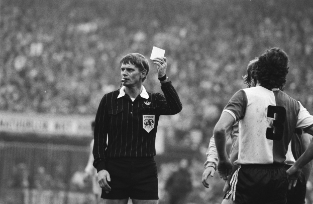 Schwarzes und weißes Bild eines Schiedsrichters mit einer roten Karte, mit einer Menge von Menschen in Fußballuniformen hinter einem Zaun.