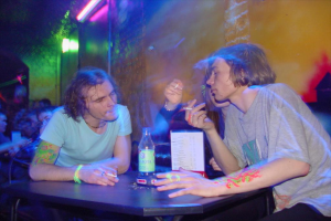 Ein Tisch mit einer Flasche, einer Speisekarte und einem Aschenbecher in der Mitte einer Indoor-Einrichtung, mit zwei Männern und einer Frau, die rauchen.