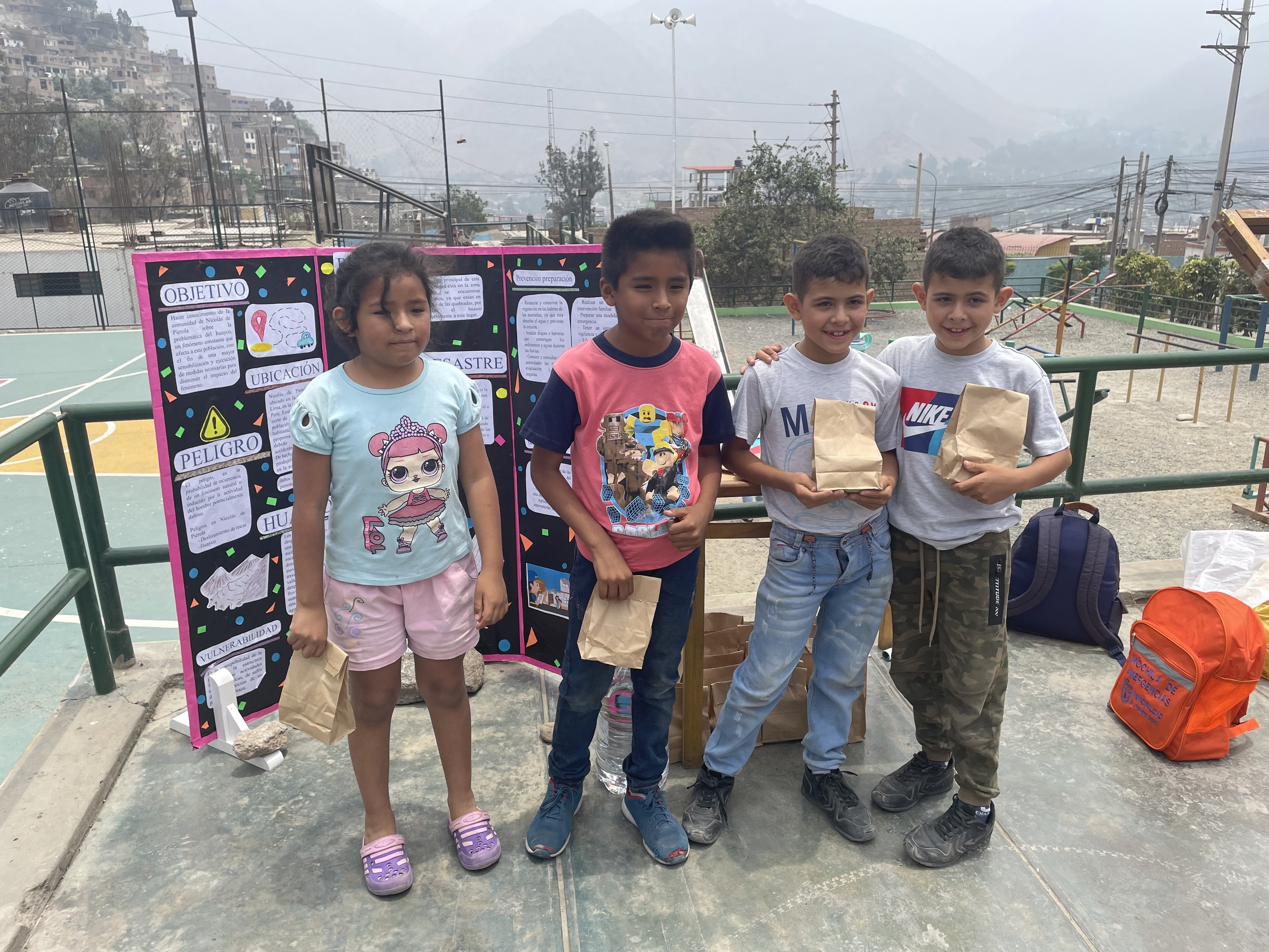 Gruppe von Kindern auf einem Basketballplatz mit Papiertüten in den Händen, mit Gegenständen auf der rechten Seite und einer Tafel mit Text auf der linken Seite, vor einem Hintergrund aus Zäunen, Pfosten, Straßenlaternen, Bäumen, Gebäuden, Hügeln und Himmel.