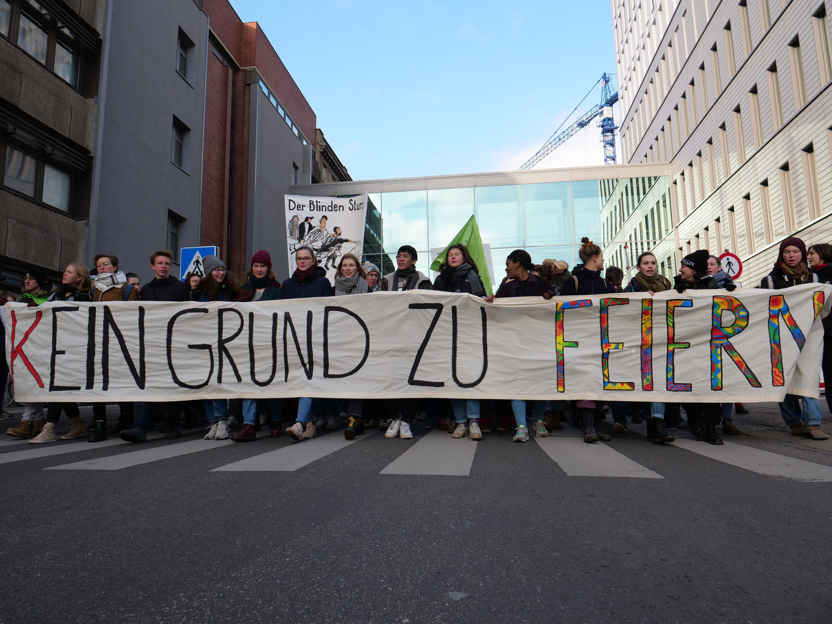 Demonstranten mit einem Banner mit der Aufschrift "Kein Grund zu Feiern" gegen die deutschen Sparmaßnahmen, im Hintergrund Gebäude, ein Schild, ein weiteres Banner, ein Kran und ein bewölkter Himmel.