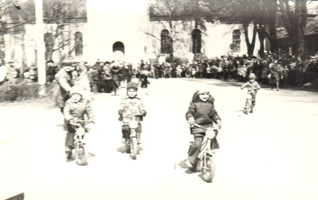 Schwarze-Weiß-Foto von Kindern auf Fahrrädern, die eine Straße mit einem Gebäude und Bäumen im Hintergrund hinunterfahren.