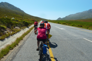 Eine Gruppe von Menschen mit Helmen auf Fahrrädern auf einer Straße, mit Pflanzen, Gras, Pfählen, Bergen und einem bewölkten Himmel im Hintergrund.