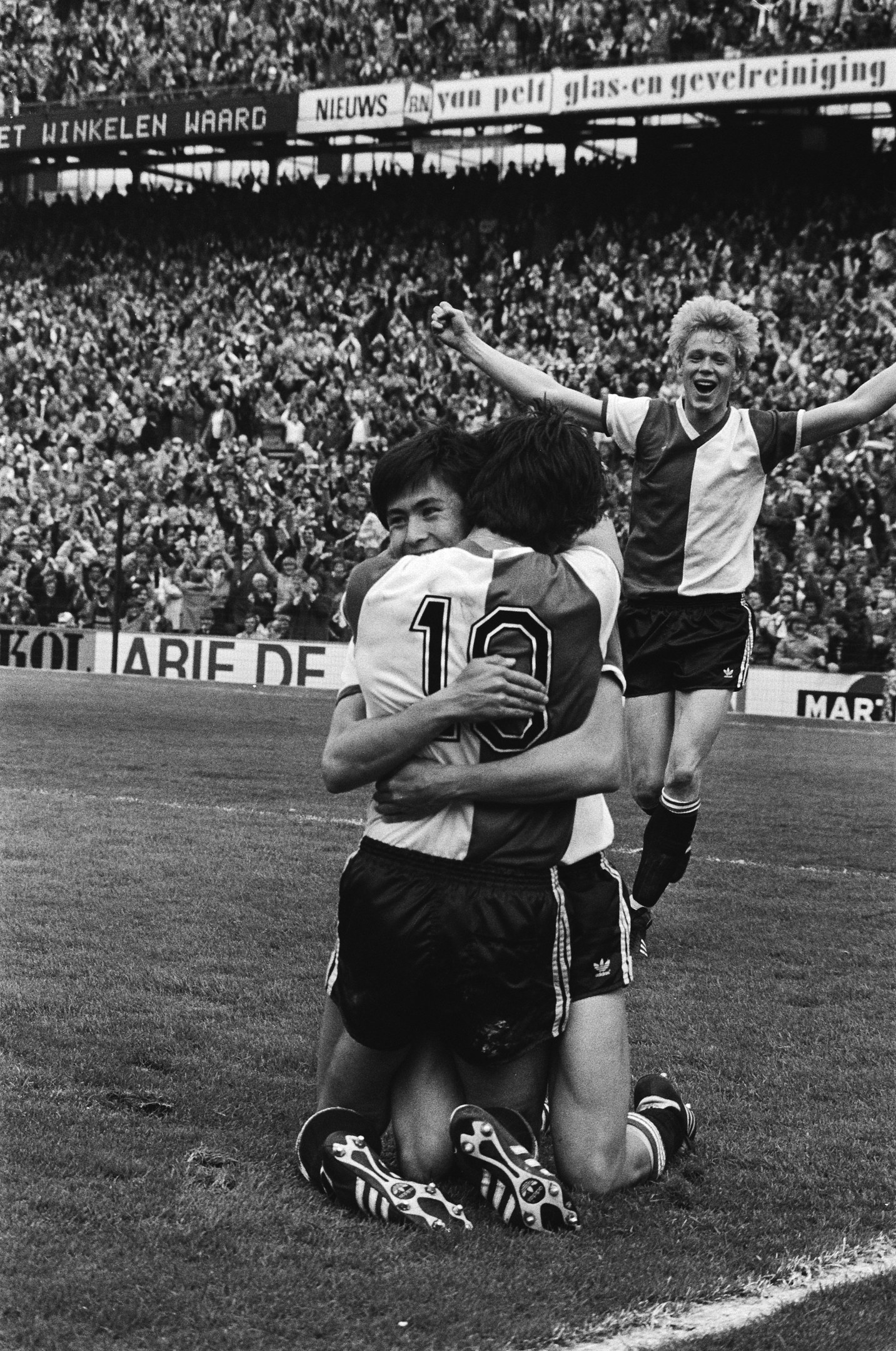 Drei Fußballer liegen sich nach einem Tor jubelnd in den Armen, mit einer jubelnden Menge und schwenkenden Fahnen im Hintergrund, alles in Schwarz-Weiß.