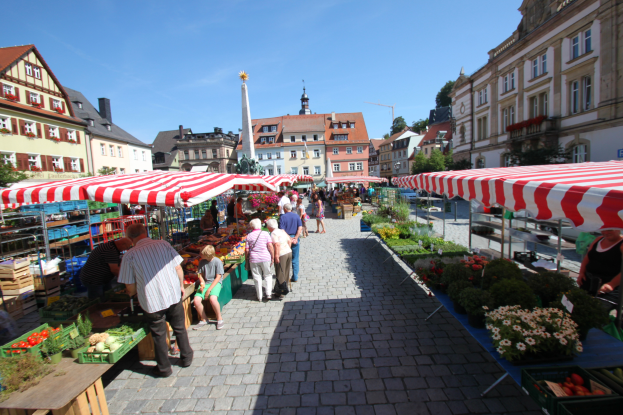 Ein belebter Markt in Heidelbergs altem Stadtkern mit Menschen, die gehen, sitzen und stehen, umgeben von Zelten mit Körben voller Gemüse, vor einer Kulisse aus Gebäuden, Bäumen und einem klaren blauen Himmel.