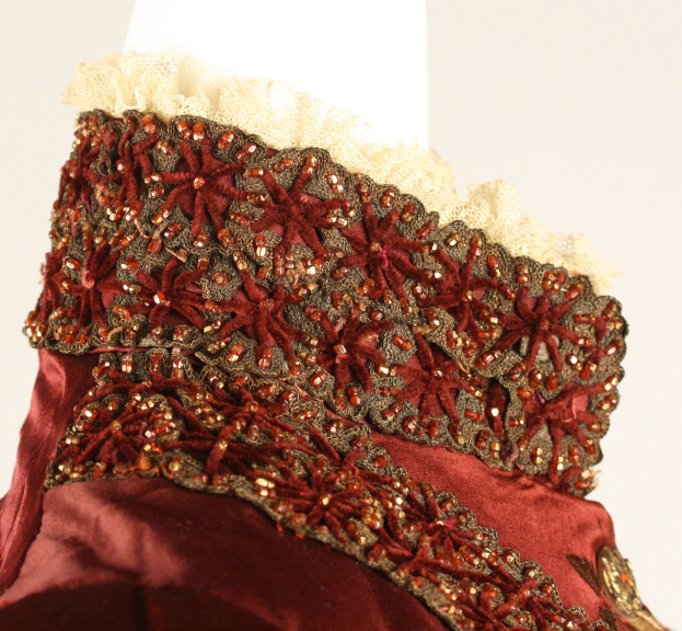 Ein Close-up eines roten und goldenen Kleides auf einer Schaufensterpuppe, das detailreiche Spitzenverzierungen auf einem weißen Hintergrund zeigt.