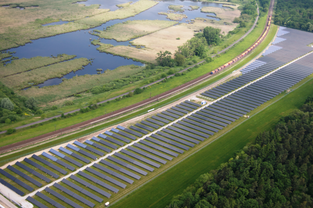 Eine Luftaufnahme eines Solarparks mit zahlreichen Solarpanelen in einem Feld, umgeben von Bäumen, Gras und Wasser, mit einem Zug, der auf einem Bahngleis in der Nähe fährt.