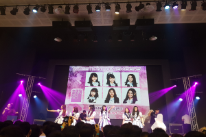 Eine Gruppe von Menschen, die auf einer hell erleuchteten Bühne performen, mit einer Menge darunter, einem Sänger mit Mikrofon und einem Musiker, der ein Instrument spielt, während ein großer Bildschirm Bilder der K-Pop-Gruppe BK48 anzeigt.