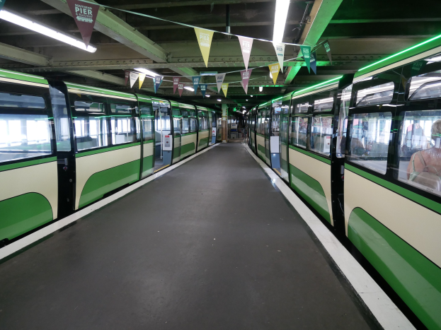 Grüner und weißer Zug fährt auf Schienen neben einem Bahnsteig mit Passagieren im Inneren, unter einer Decke mit Lampen und dekorativen Flaggen an einem Bahnhof in München, Deutschland.