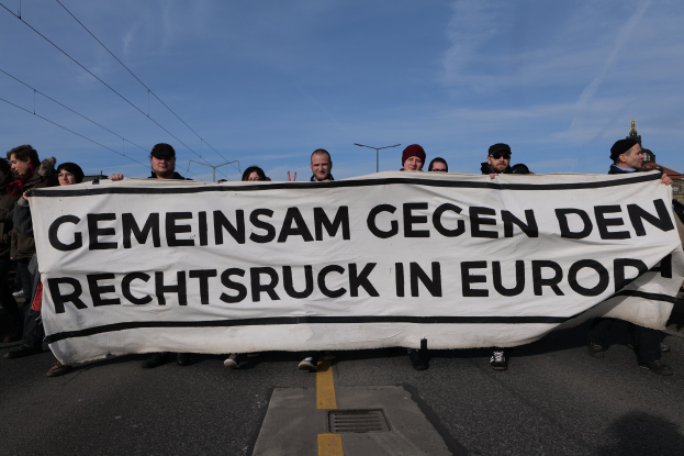 Eine Gruppe von Menschen hält ein Transparent mit der Aufschrift "Gemeinsam gegen den Rechtsruck in Europa" auf einer Straße mit Straßeninfrastruktur und Gebäuden im Hintergrund.