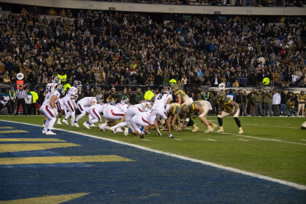 Ein Fussballspiel wird in einem Stadion gespielt, wobei Spieler in Sportkleidung und Helmen auf dem Feld sind, während Zuschauer und verschiedene Gegenstände im Hintergrund zu sehen sind.